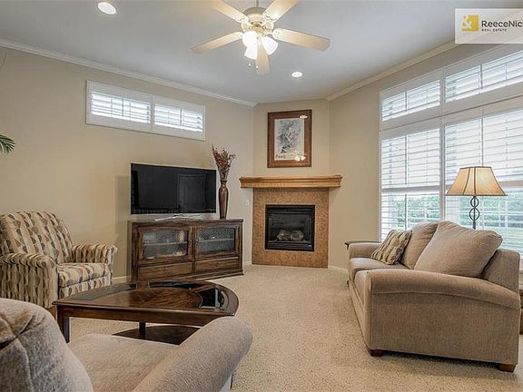 Warm and cozy living room with carpet, large windows looking out to the pond, a gas fireplace and ceiling fan.
