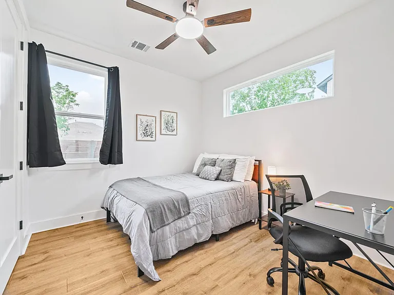 Furnished bedroom with bed, desk workspace, ceiling fan, and windows with coverings for natural light and privacy.