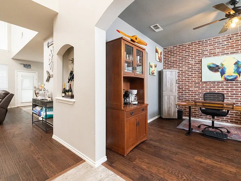 Dedicated office space with a desk, ergonomic chair, and coffee cabinet. Brick accent wall and ceiling fan create a cozy and inspiring work environment.