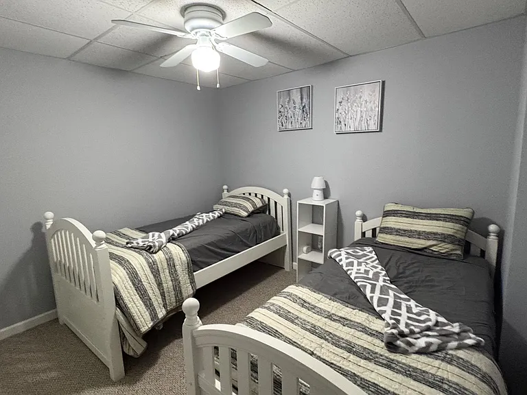 Furnished bedroom with two twin beds, a shared nightstand, ceiling fan lighting, and neutral walls for easy setup.