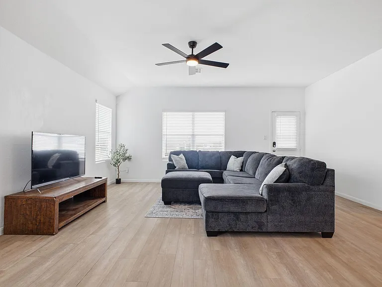 Open living room with an L-shaped sectional, ceiling fan, and space for a media setup, plus wide-plank flooring.