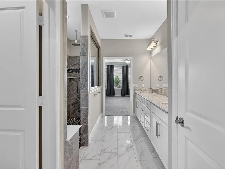 Spacious bathroom featuring white cabinetry, dual sinks with stone countertops, and a large shower with dark tile and pebble flooring