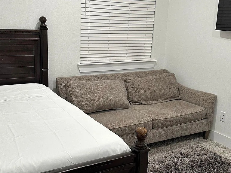 Furnished bedroom with a bed, loveseat seating area, wall-mounted TV, and a window with blinds for privacy.