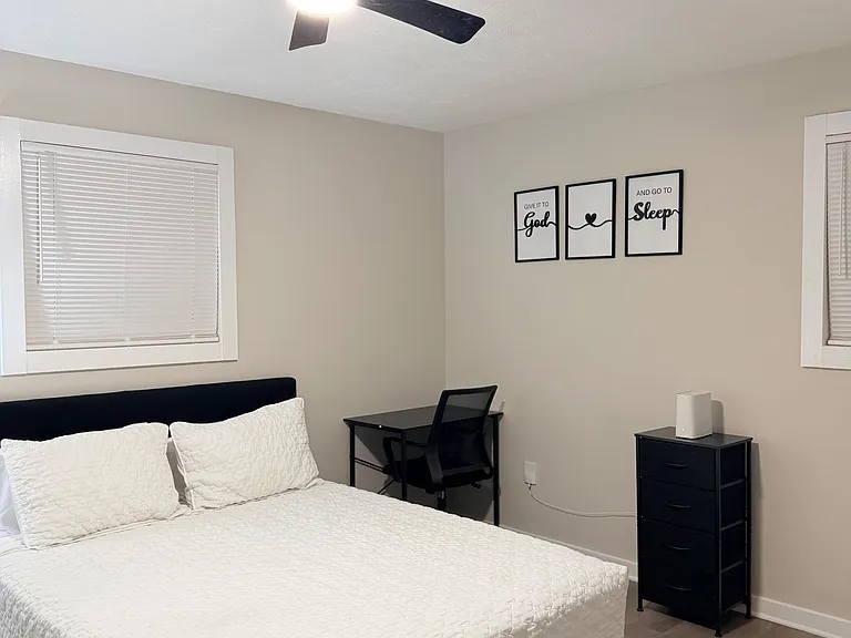 Furnished bedroom with a full bed, desk setup, dresser storage, and a ceiling fan with light plus windows with blinds.