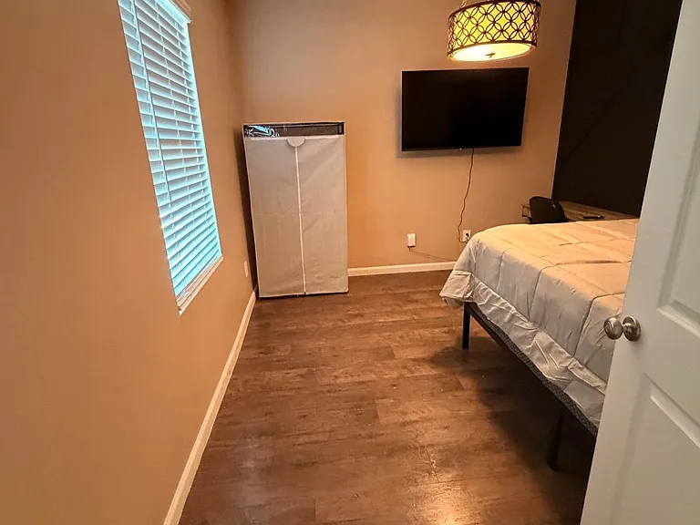 Private bedroom with wood-look flooring, a large window with blinds, mounted TV, and space for a bed and storage.