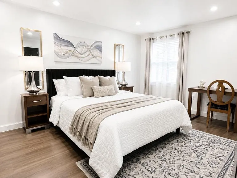 Spacious bedroom with a queen-size bed, nightstands with lamps, a dedicated desk area, and a window with curtains.