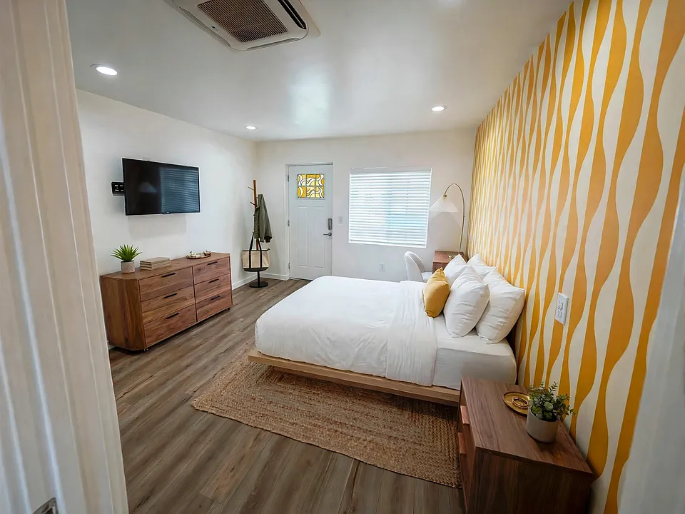 Spacious studio layout featuring custom yellow accent wall, wall-mounted TV setup, and modern vinyl plank flooring. (Furnished for display)