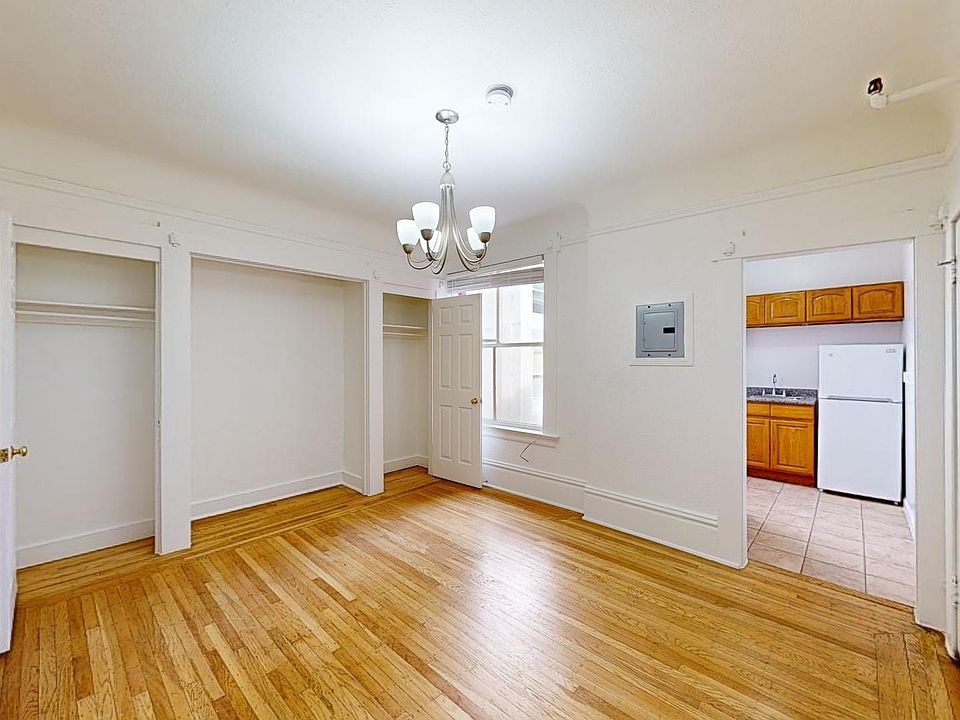 Bright, empty room with wooden flooring, walls, and a chandelier. Open closets, a window, and a view into a kitchen with wooden cabinets.