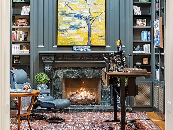 The library office  is adorned with tasteful finishes and decor. The room features rows of  built-in bookshelves, a cozy fireplace, and plenty of natural light. This handsome library office combines c