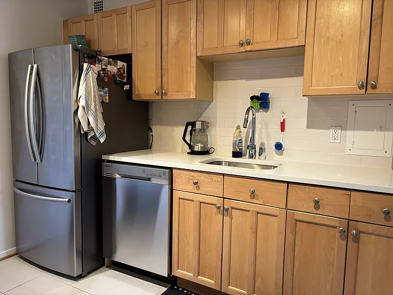 One side of the kitchen, with ample cabinets, dishwasher, fridge/freezer, sink with removable faucet head, and electric kettle. Ample cleaning supplies under the sink.