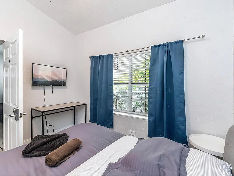 Private bedroom with a large window, blackout curtains, wall-mounted TV, and a compact desk area for work or study.