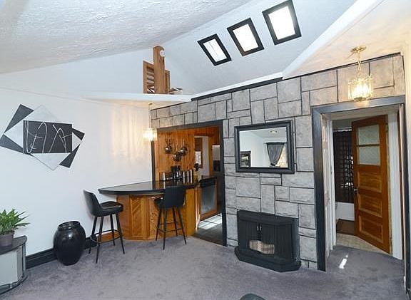 Spacious living room with faux fireplace and stone wall opens to kitchen.