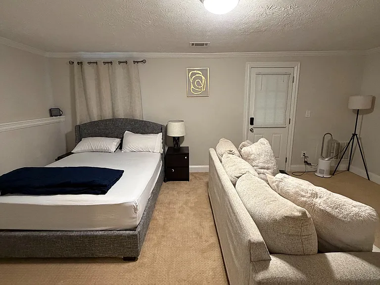 Furnished bedroom with a full bed, nightstand lighting, and space for seating, plus a covered window and direct exterior door