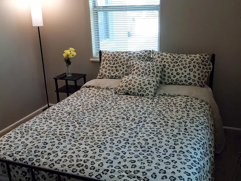 Private bedroom features a full-size bed, side table, and two windows with blinds for natural light and privacy.