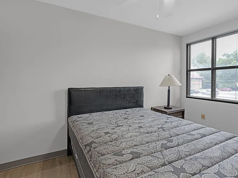 Furnished bedroom featuring a comfortable bed, nightstand with lamp, and a large window with blinds for natural light.