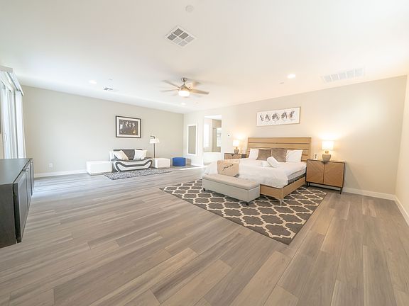 A large bedroom featuring a comfortable bed, stylish furniture, and ample natural light. The neutral tones and wooden accents create a cozy yet modern atmosphere.