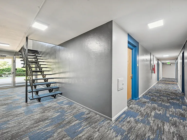Contemporary apartment hallway featuring modern blue and gray carpeting, sleek metal staircase, and bright natural lighting.