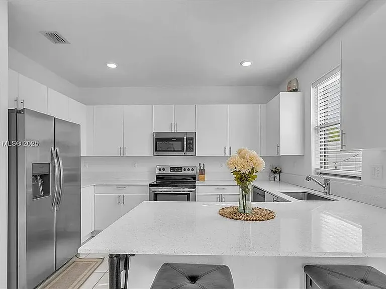 Modern all-white kitchen with quartz counters, stainless steel appliances, and ample natural light through a window above the