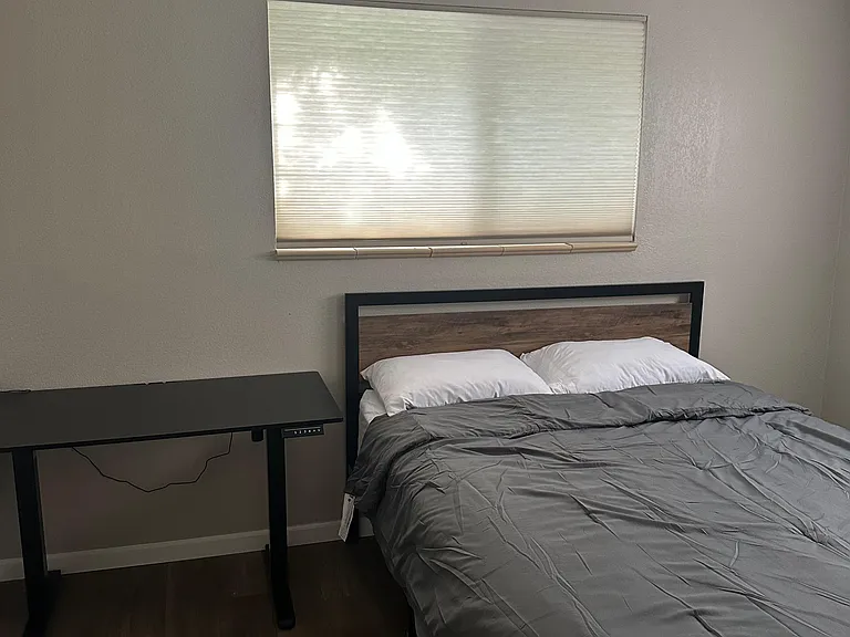 Private bedroom with a comfortable bed, nightstand/desk space, and a window with blinds for light and privacy.