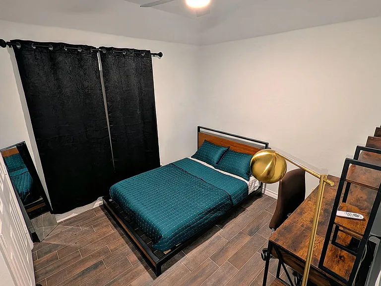 Furnished bedroom with a full bed, tile flooring, blackout curtains, and a compact workspace with shelving.
