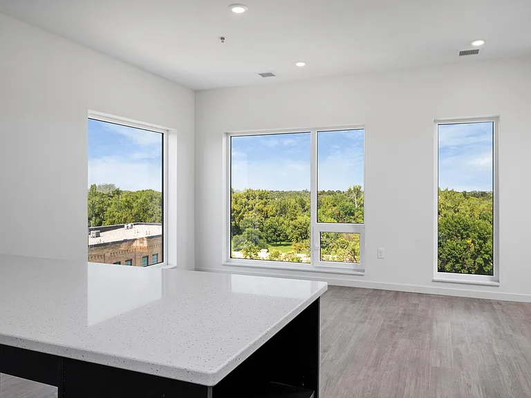Bright and spacious apartment interior with large windows offering a stunning view of lush greenery, complemented by sleek hardwood floors and a modern white countertop. Bright and spacious apartment interior with large windows offering a stunning view of