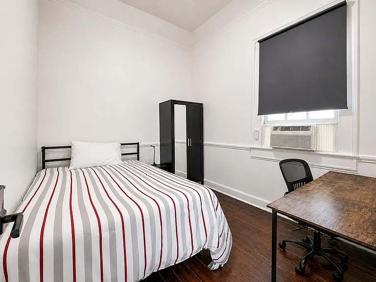 Private furnished bedroom with a full bed, wardrobe storage, and a dedicated desk area, plus window A/C and dark wood floors.