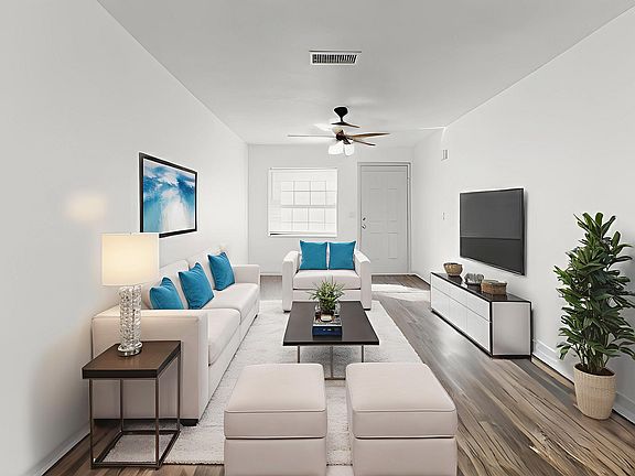 Bright and inviting living room with sleek modern decor, featuring white sofas, blue accent pillows, and a stylish coffee table. A flat-screen TV is mounted on the wall, complimented by elegant hardwood flooring and lush green plant for a touch of nature.