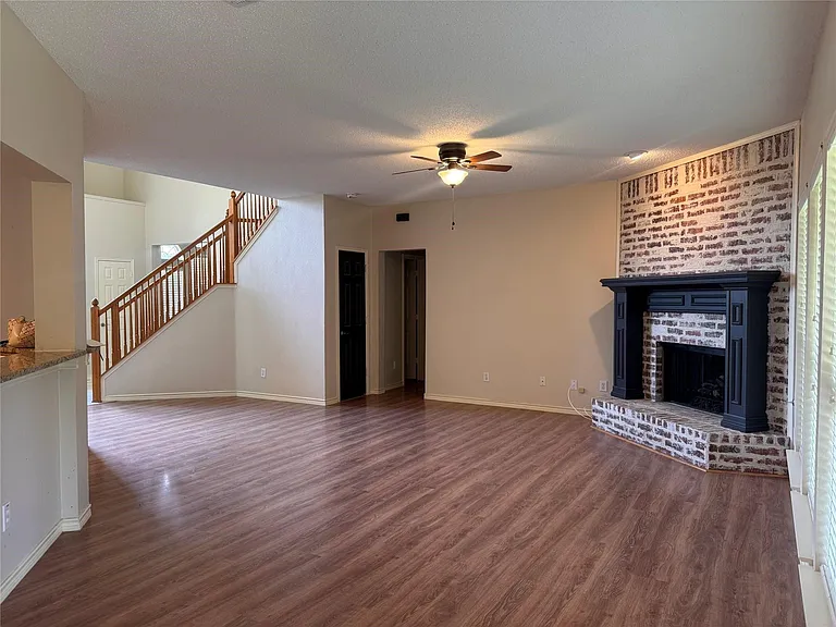 Spacious room featuring wood-finish flooring, a white-washed brick fireplace with a black mantel, a ceiling fan, and a wood staircase with balusters