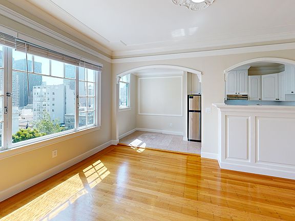 Sunlit living room with a large window, wood-like flooring, and an entrance to the kitchen