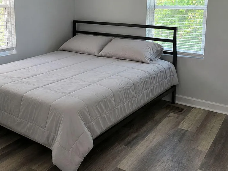 Furnished bedroom with a full-size bed, ceiling fan, and two windows with blinds overlooking greenery.