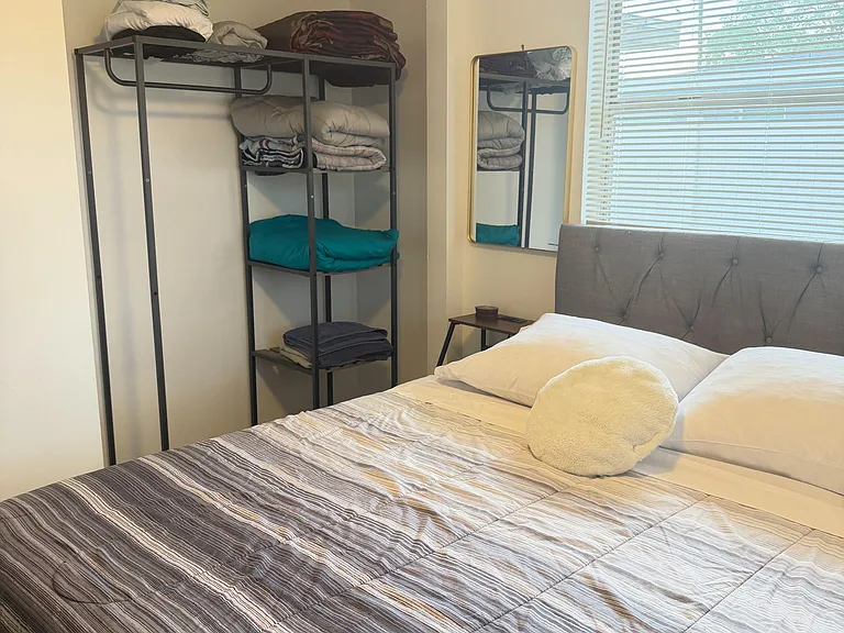 Furnished bedroom with a comfortable bed, window with blinds, and open closet storage with shelving for linens and essentials