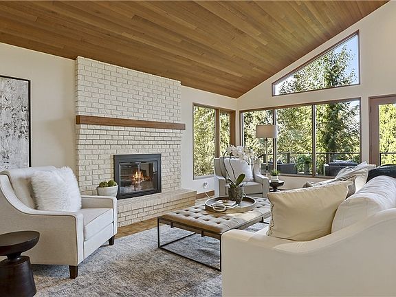 Light-filled living room with brick fireplace, pristine vaulted cedar ceilings, intricate hardwood floors and door to  deck offering peaceful views of mountains, trees and rolling meadow.