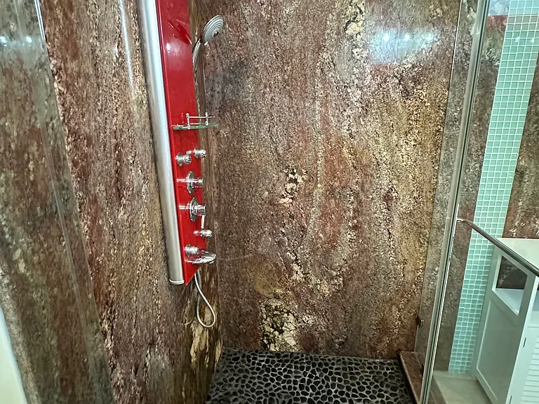 Spa-style shower featuring granite slab walls, river rock flooring, a rain showerhead, handheld sprayer, and body jets.