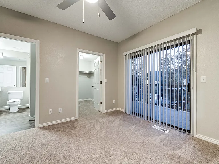 Bright bedroom with modern ceiling fan, showcasing a spacious walk-in closet, adjoined elegant bathroom, and floor-to-ceiling glass doors leading to a scenic balcony view.