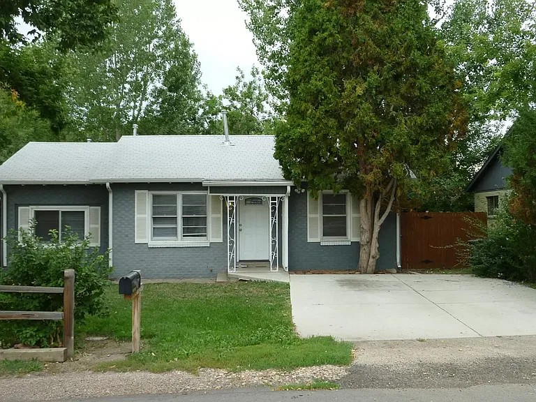 Quaint single-story gray house with a white roof, front porch, and lush green surroundings.
