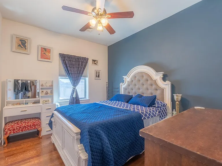 Furnished bedroom with a queen bed, ceiling fan hardwood floors, and a window with curtains plus a vanity and dresser storage