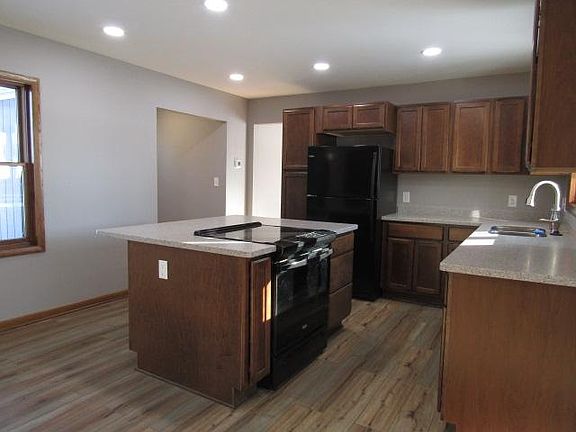View of entry hall and kitchen, which comes equipped with black appliances, eat-at island with built in stove, wood cabinets, man-made granite counter tops, faux wood laminate flooring, spotlights, no window treatments.