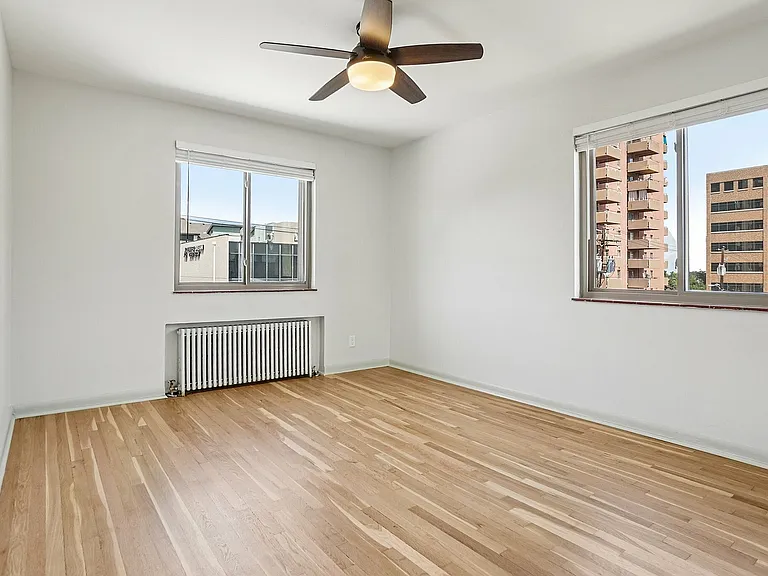 Bright and spacious bedroom with hardwood floors, large windows offering city views, and a ceiling fan for comfort.