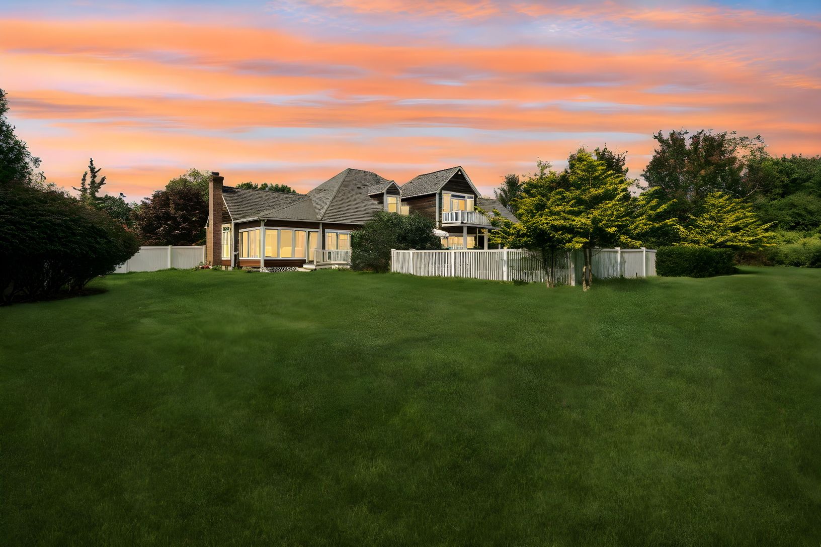 A twilight view of the home’s rear elevation, featuring the fenced backyard, chimney, and sunroom.