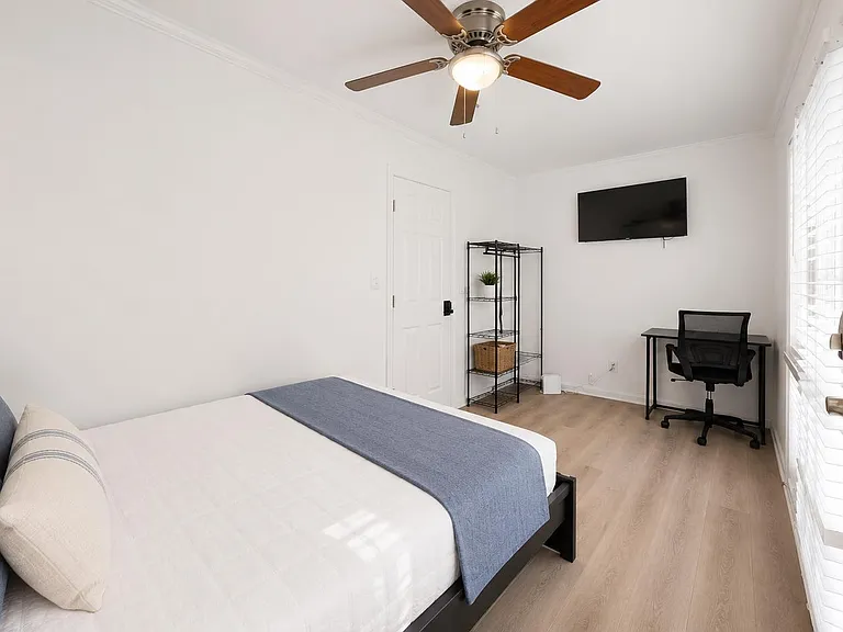 Private bedroom with a full bed, work desk, wall-mounted TV, and large windows with blinds for natural light.