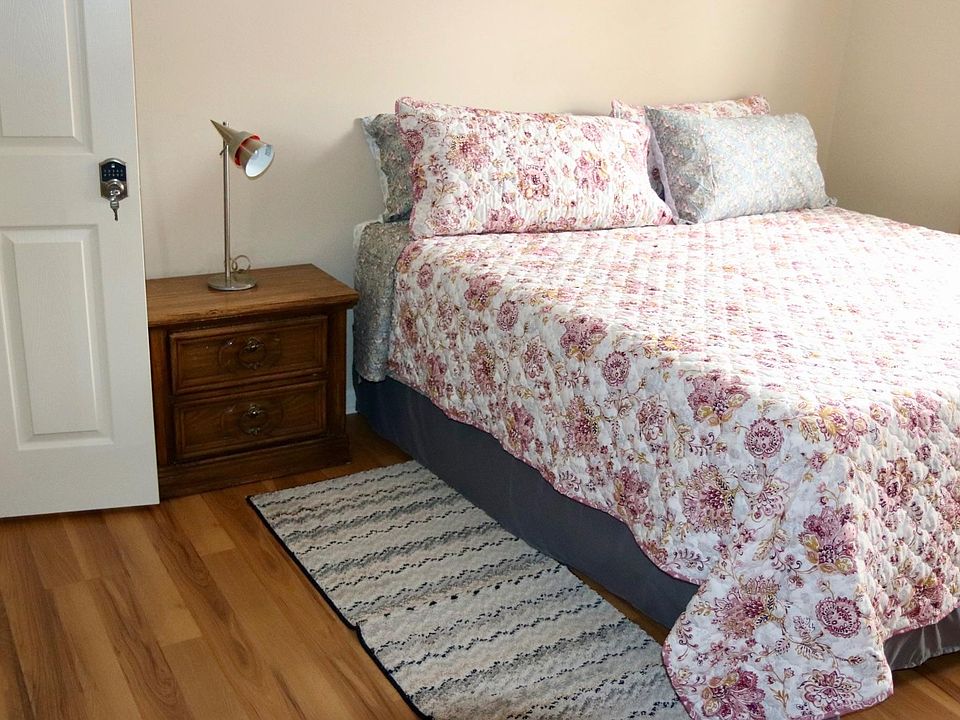 Furnished bedroom with a full-size bed, nightstand and reading lamp, plus a window and hardwood flooring.