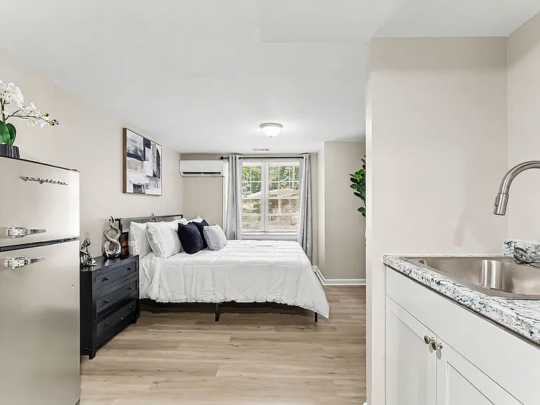 Efficient studio-style bedroom with a dedicated sink area, mini fridge, dresser storage, and a large window with blinds.