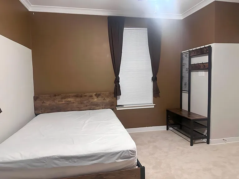 Furnished bedroom with a full bed, window with blinds and curtains, and a hall tree/bench for storage and organization.