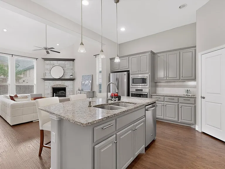 A modern kitchen featuring a kitchen island with dual sink, stainless steel appliances, warm hardwood floor and decorative lighting, creating a stylish and inviting atmosphere.