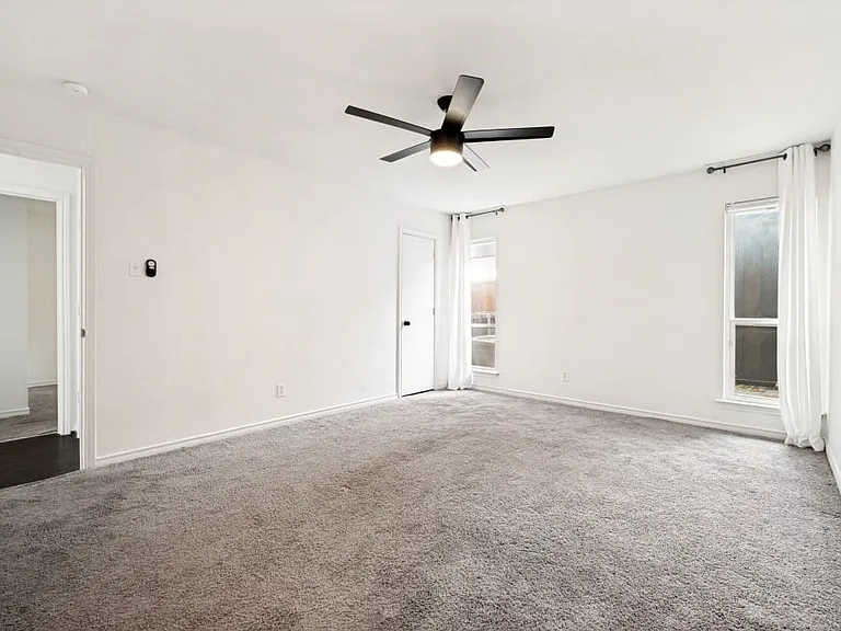 Light gray carpeted room with white walls, a black ceiling fan with integrated lighting, and two vertical windows with white curtains