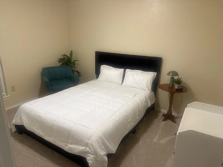 Furnished bedroom featuring a full bed, nightstand with lamp, and a comfy accent chair with space for a small desk.