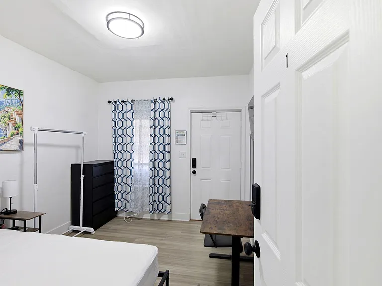 Furnished bedroom with a bed, dresser, desk, and window with curtains, offering a practical layout for work and rest.