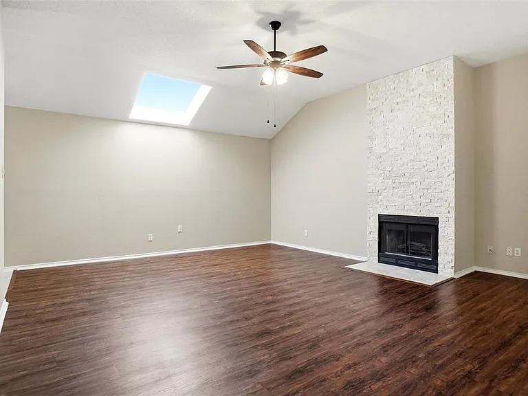 Spacious living area featuring a vaulted ceiling with a skylight, wood-finish flooring, a ceiling fan, and a fireplace with a stacked stone facade and white hearth