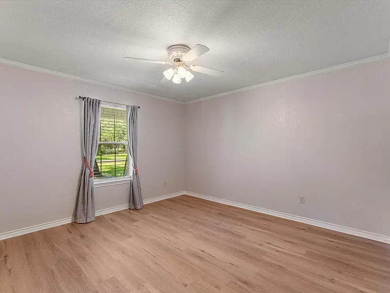 Spacious room featuring wood-finish flooring, a ceiling fan with integrated lighting, a window with exterior views, crown molding, and white baseboards