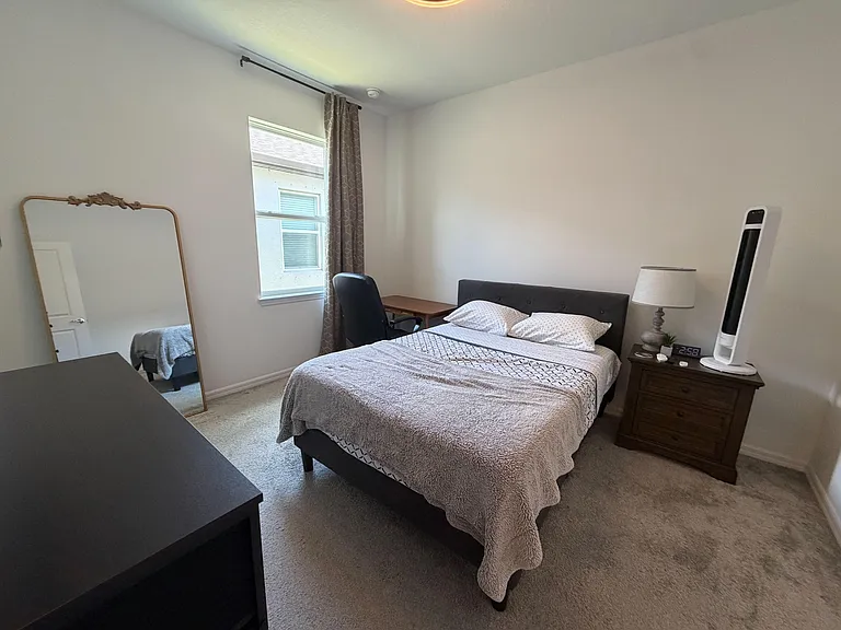 Furnished bedroom with a full bed, nightstand, and dedicated desk space, plus a window with curtains and plush carpet.